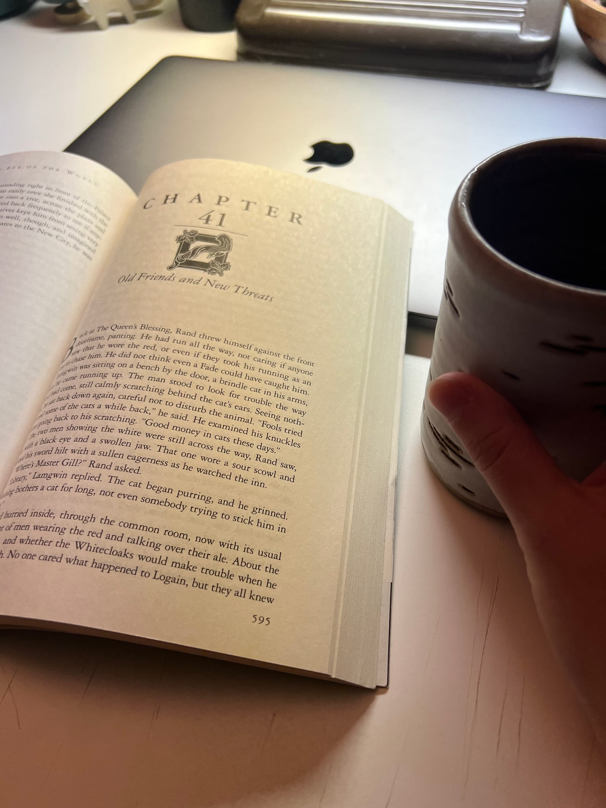 A paperback copy of The Eye of The World by Robert Jordan sits open on a desk with a hand and a cup of coffee.