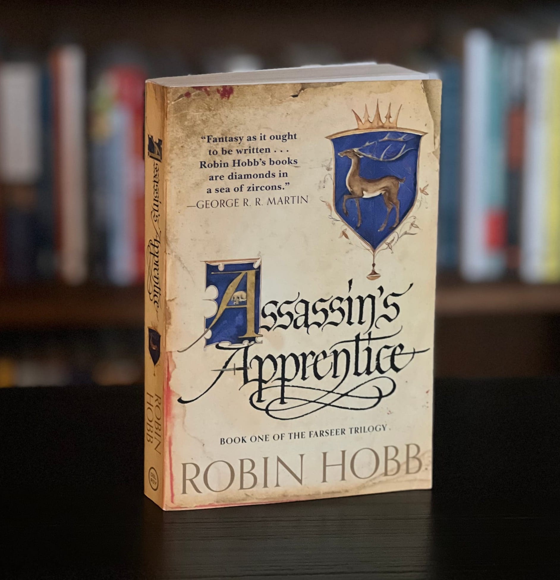 A paperback copy of Assassin's Apprentice by Robin Hobb sits on a table in front of a stuffed bookcase.