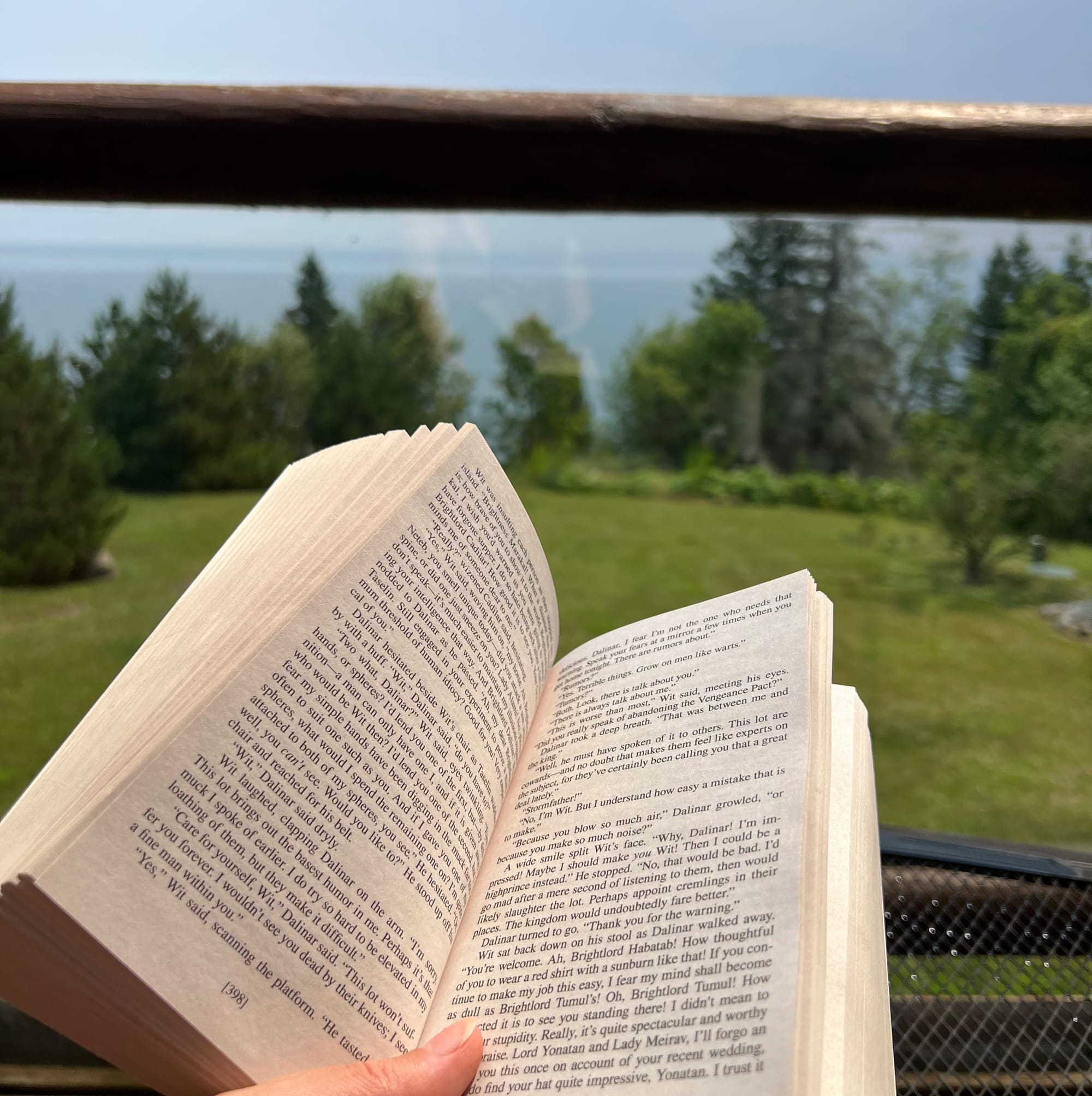 A hand holding open a paperback copy of The Way of Kings by Brandon Sanderson next to evergreen trees and Lake Superior.