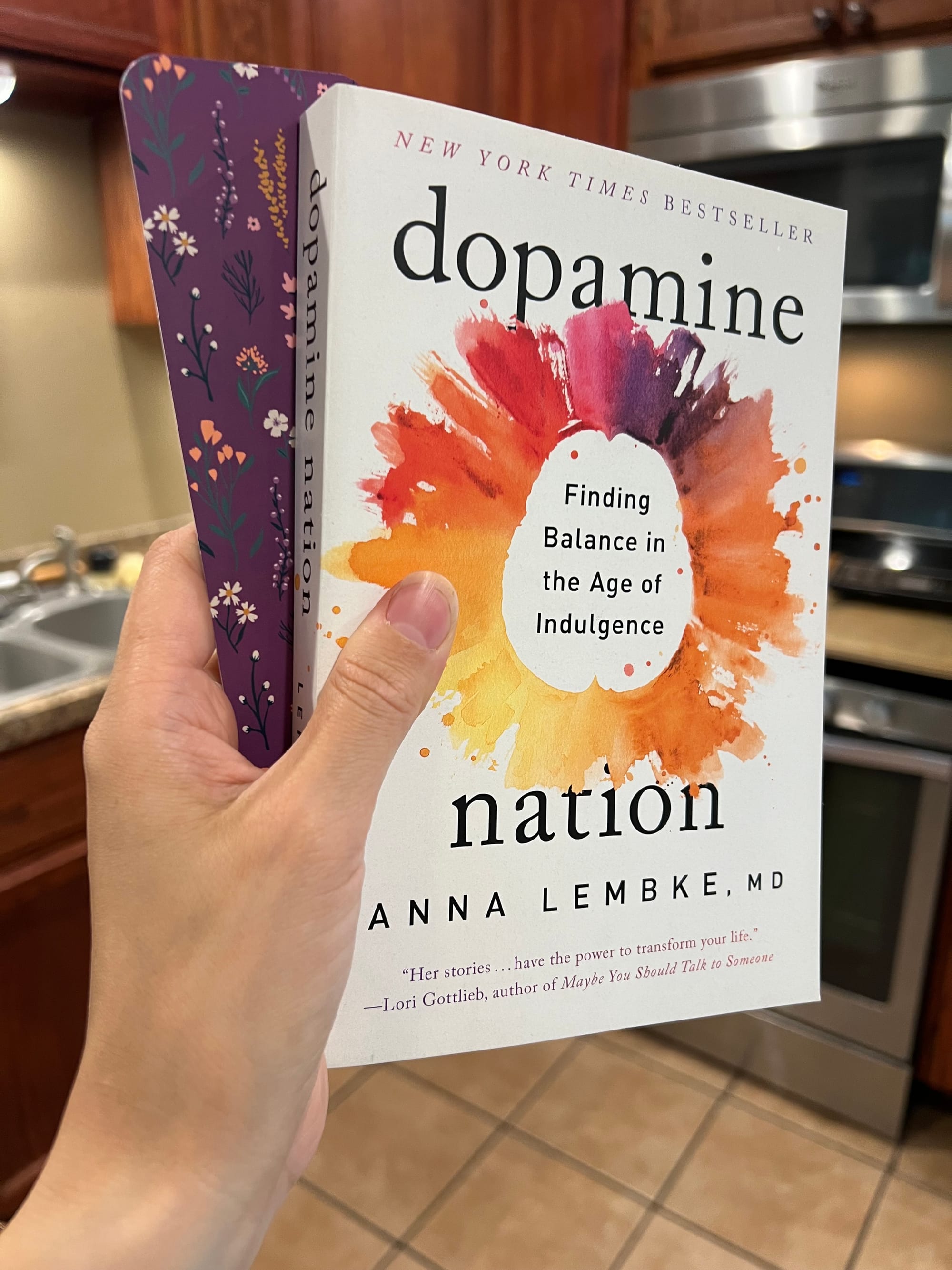 A hand holding a paperback copy of Dopamine Nation by Anna Lembke, MD in a clean kitchen.