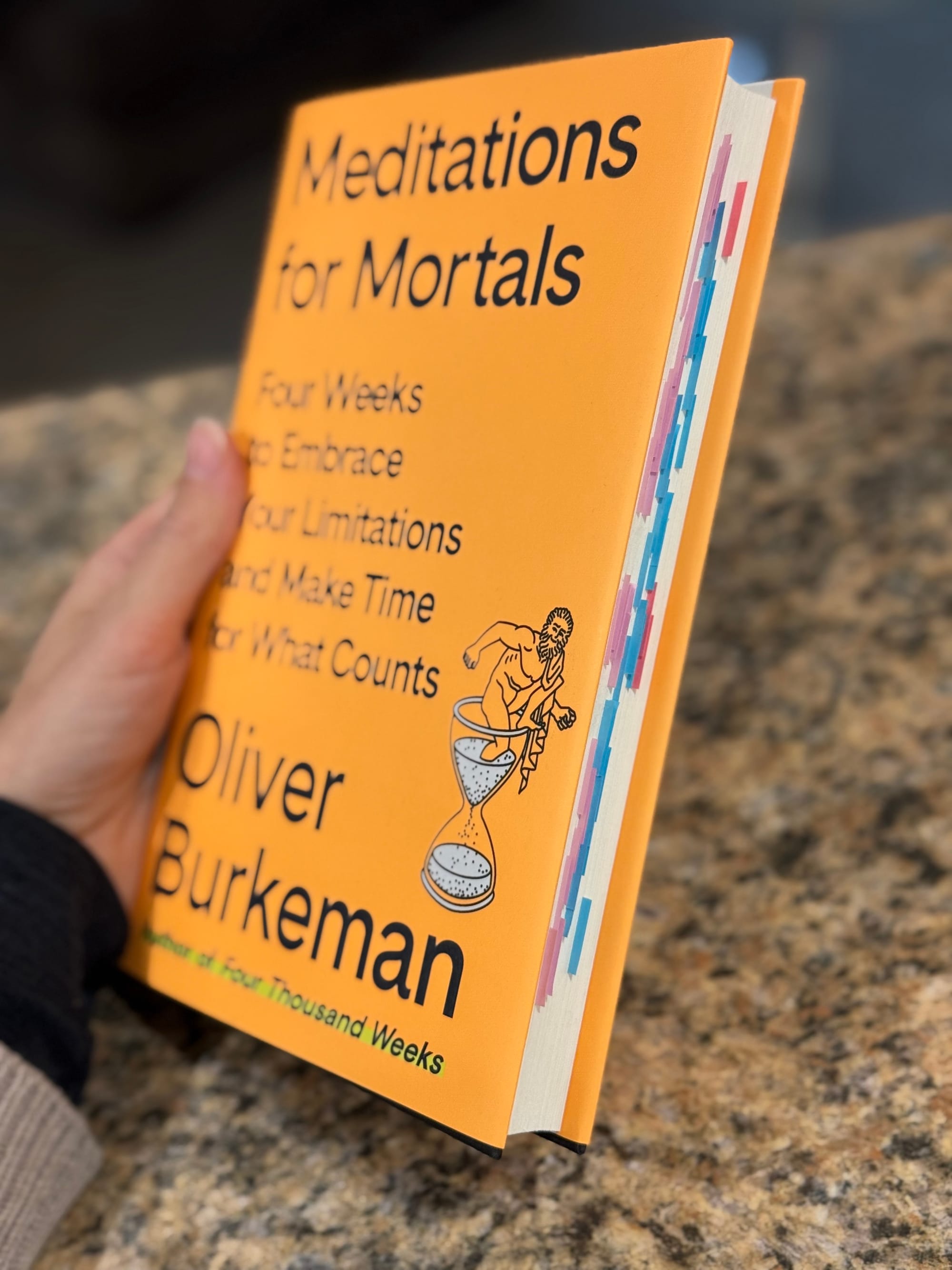 A hand holding a hardcover copy of Meditations For Mortals by Oliver Burkeman showing the annotated sticky notes on the side.