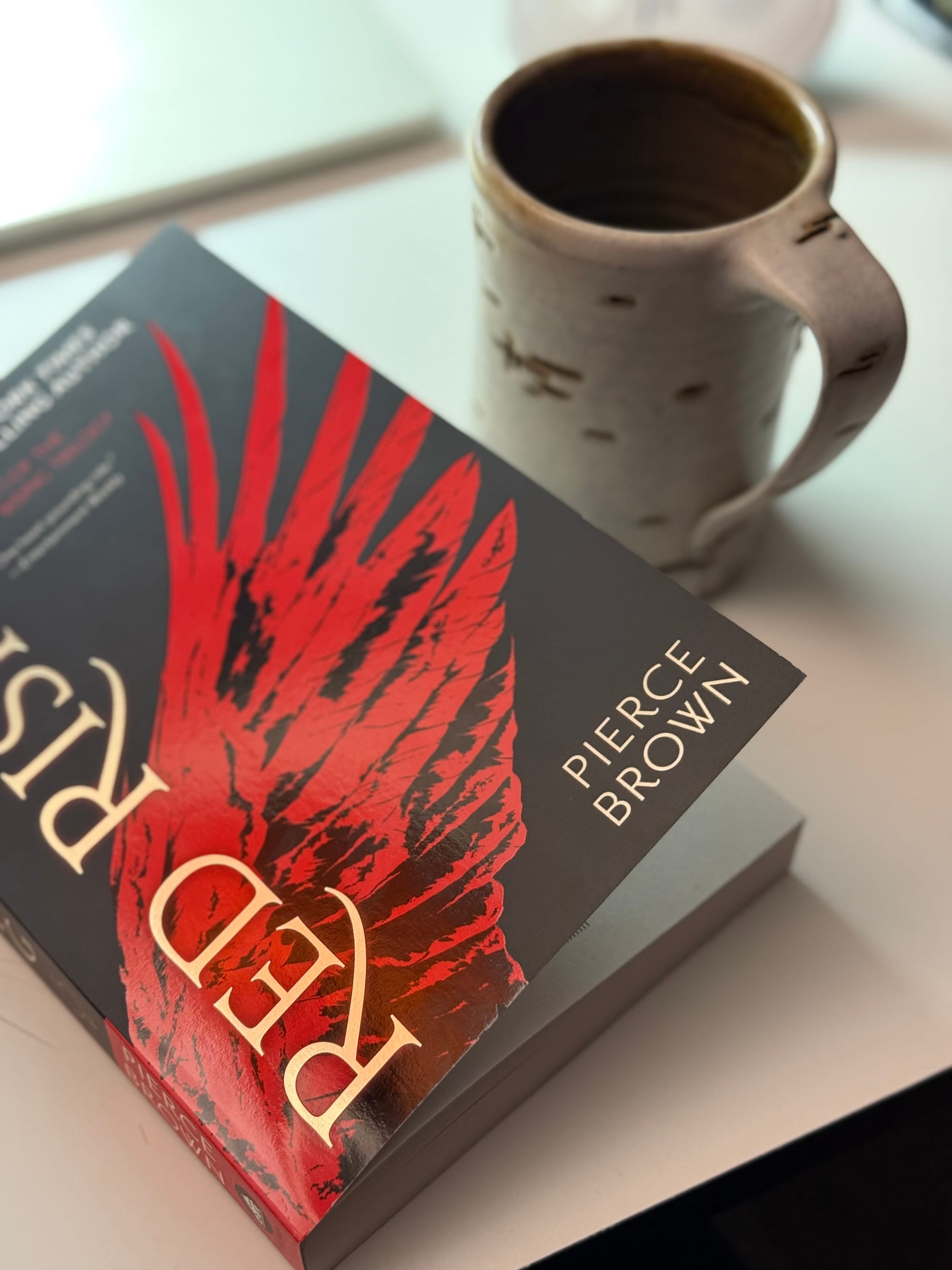 A paperback copy of Red Rising by Pierce Brown sits on a desk next to a white coffee mug.