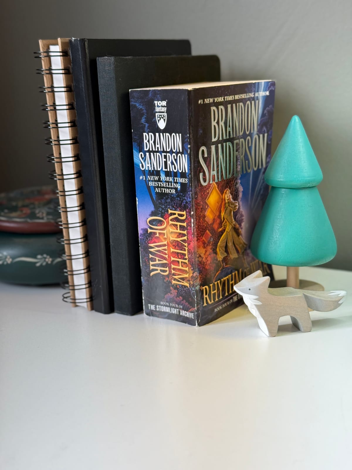 A paperback copy of Rhythm of War by Brandon Sanderson sits on a white desk next to various notebooks and journals.