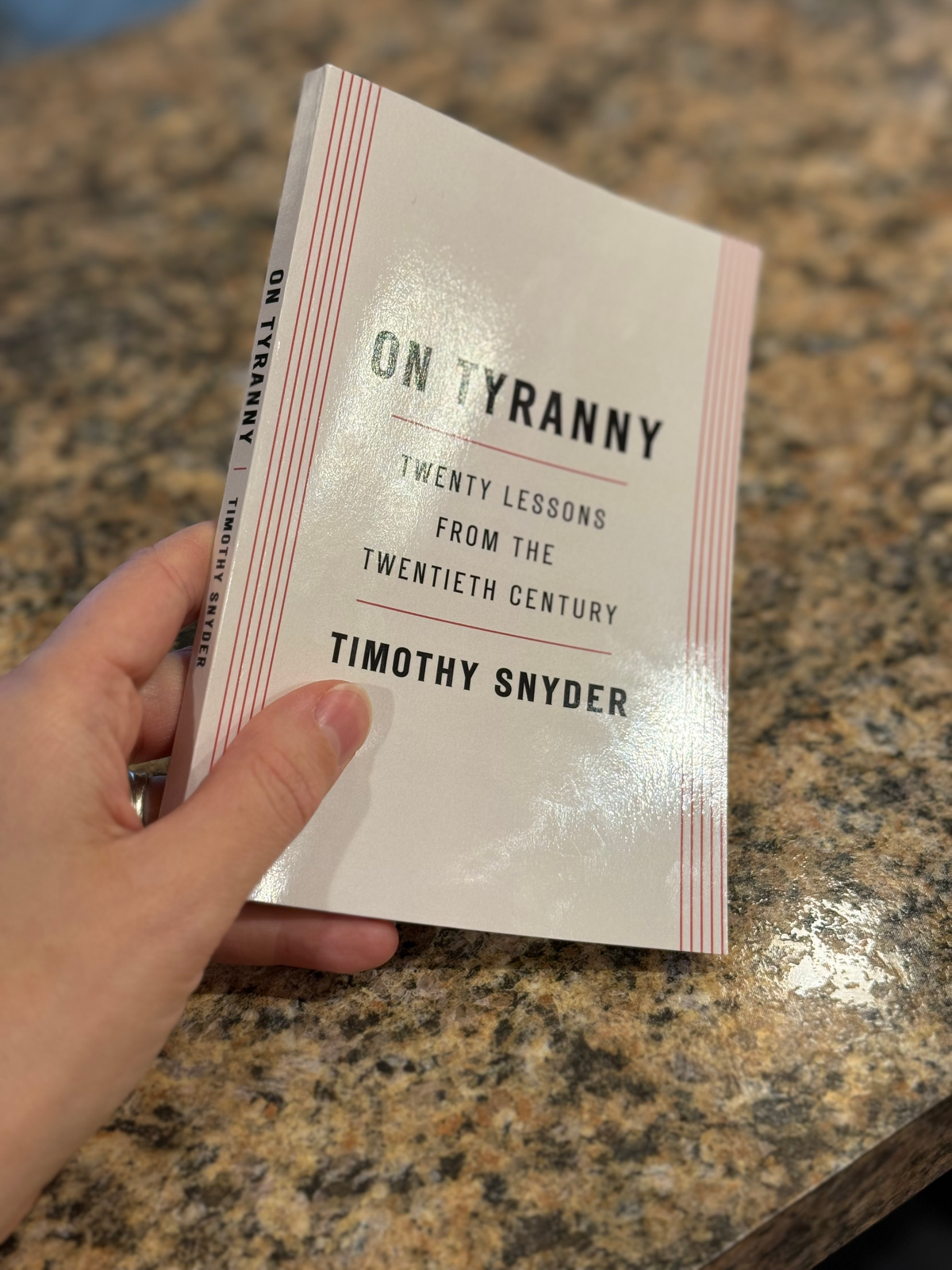 A hand holds a paperback copy of On Tyranny by Timothy Snyder on a granite countertop.