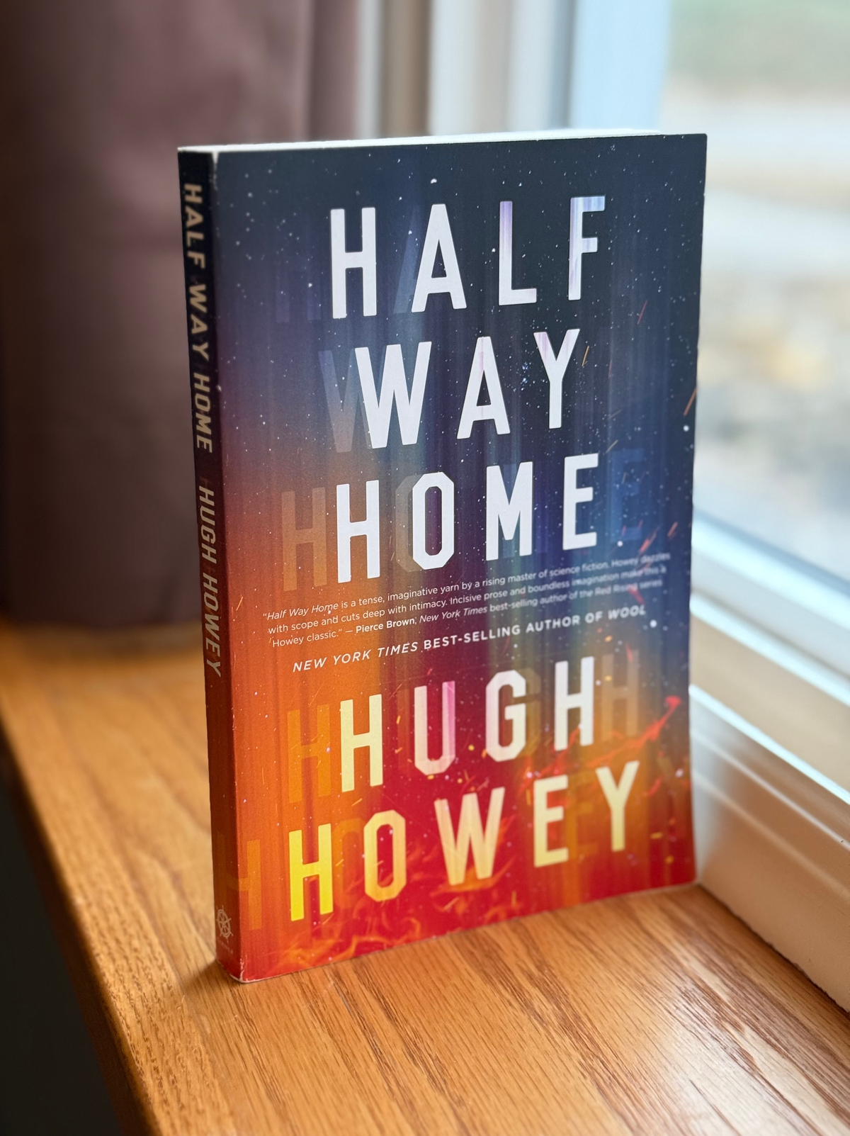 A paperback copy of Half Way Home by Hugh Howey stands on a windowsill in front of a sunny window.