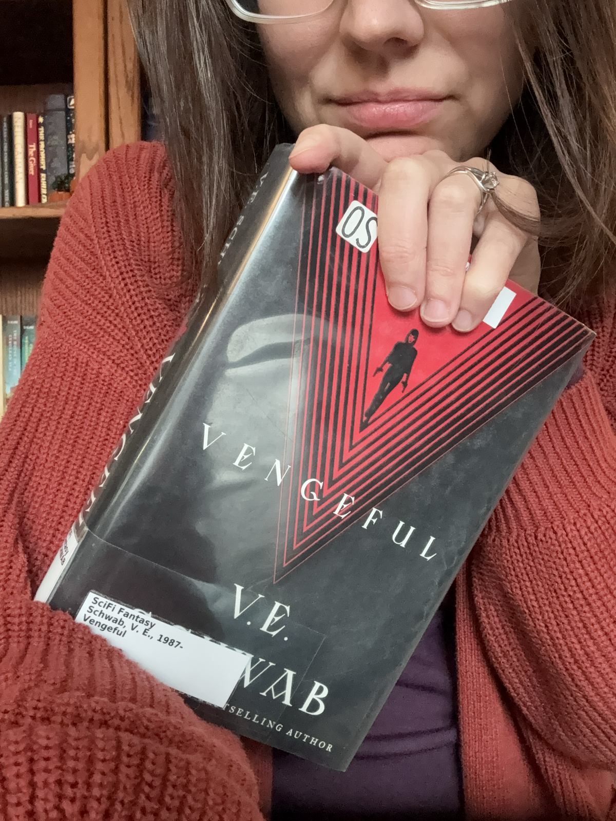 A woman holding a hardcover library copy of Vengeful by V.E. Schwab in front of a stuffed bookcase.