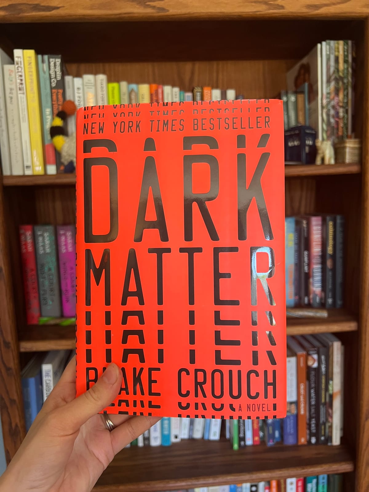 A hand holding a hardcover copy of Dark Matter by Blake Crouch in front of a stuffed bookcase.