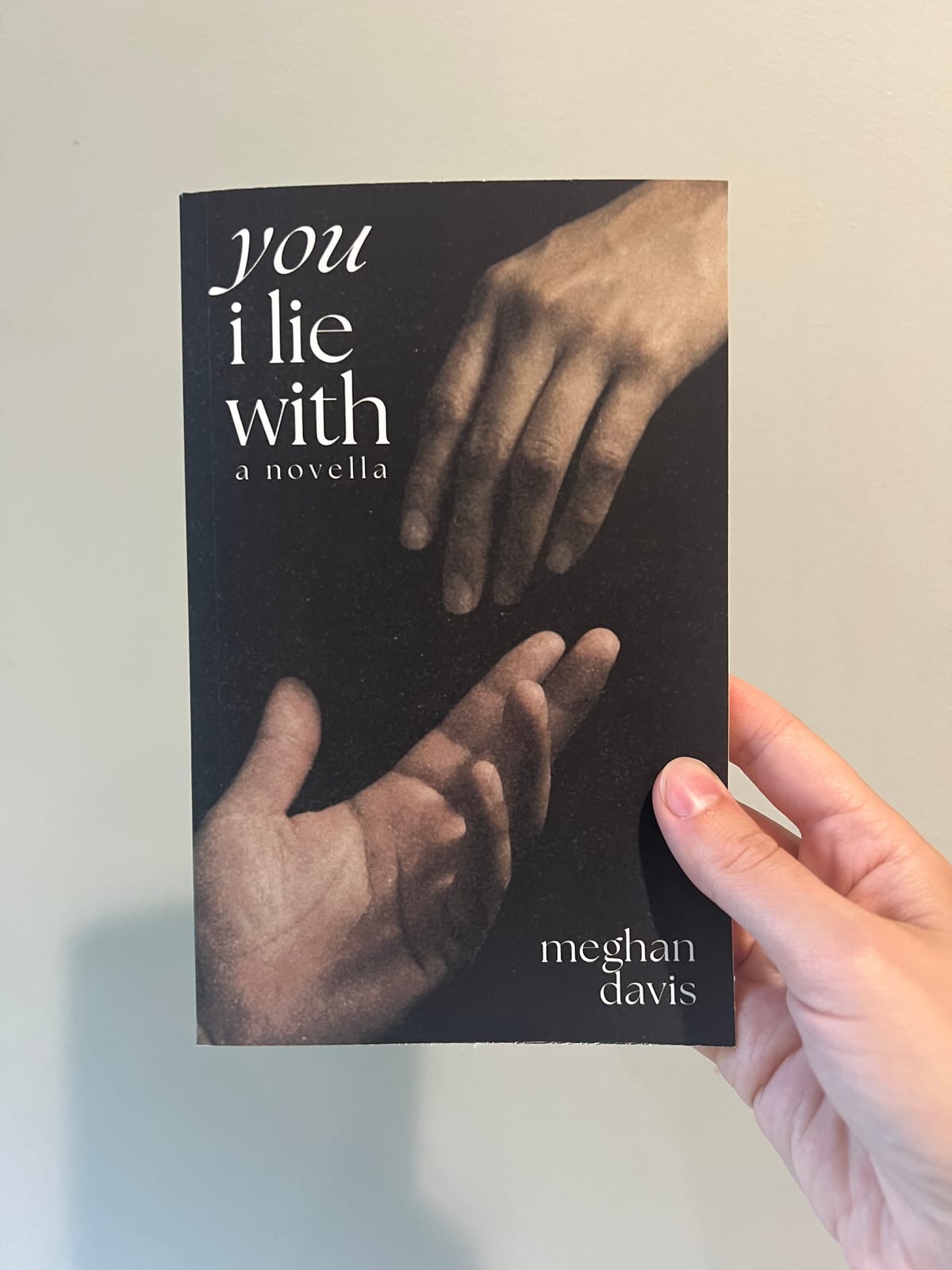 A hand holding a paperback copy of You I Lie With by Meghan Davis in front of a white wall.