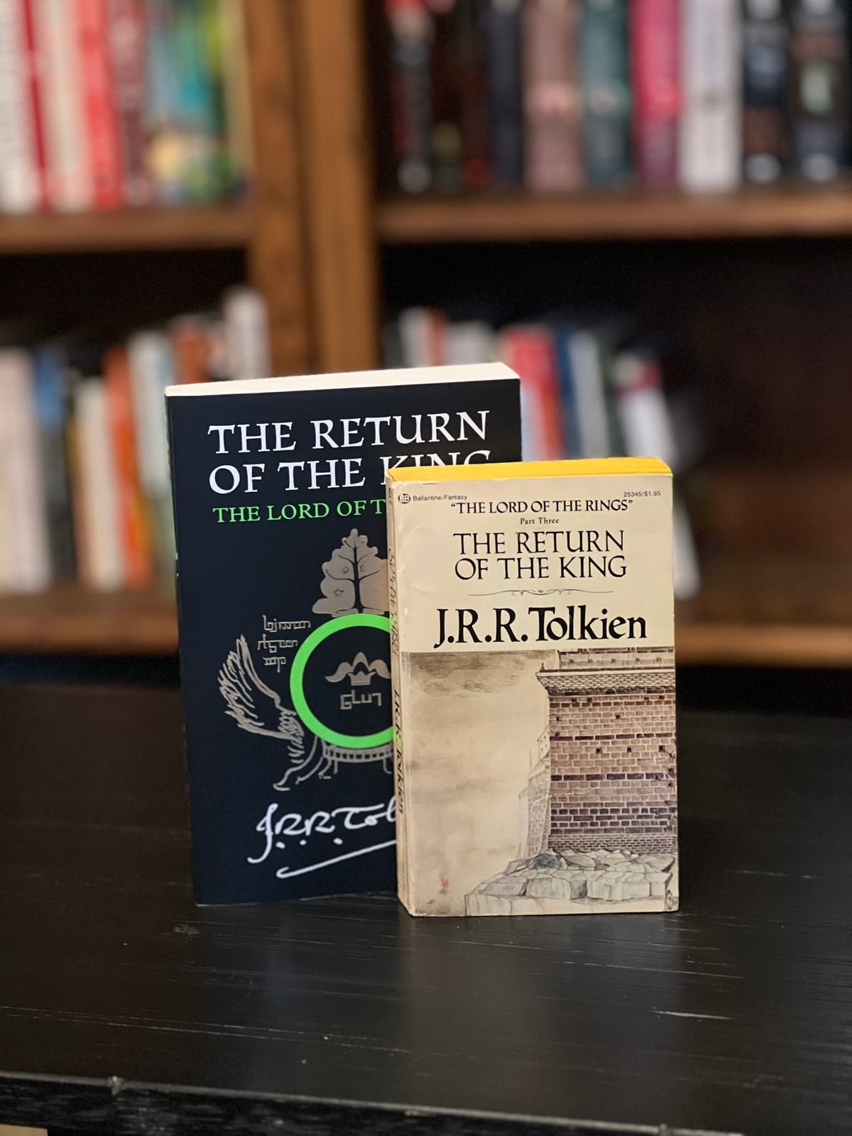 Two paperback copies of Return Of The King by J.R.R. Tolkien stand on a table in front of a stuffed bookcase.