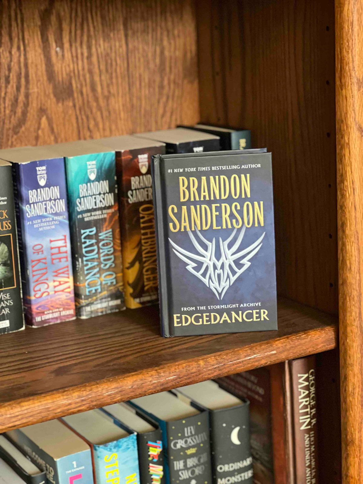 A hardcover copy of Edgedancer by Brandon Sanderson sitting on a shelf in front of a stuffed bookcase.