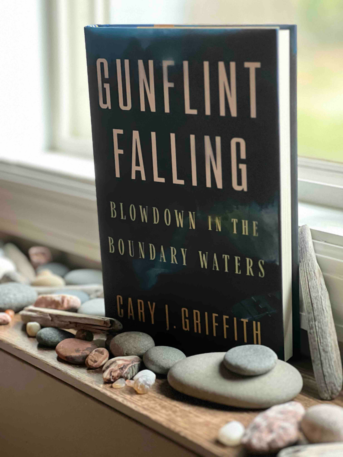 A hardcover copy of Gunflint Falling by Cary J Griffith sits on a shelf surrounded by lake rocks in front of a sunny window.