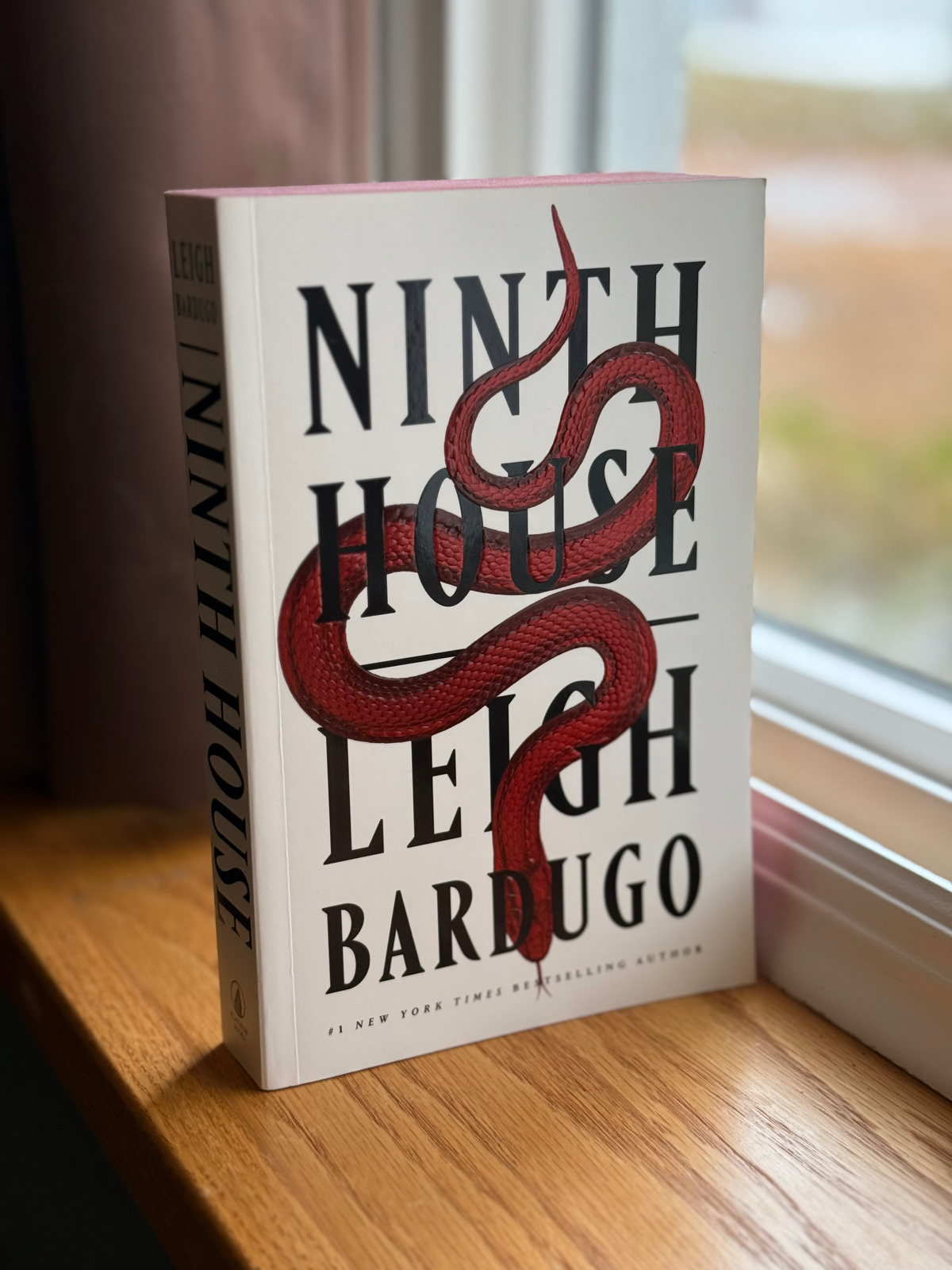 A paperback copy of Ninth House by Leigh Bardugo sits on a windowsill in front of a sunny window.