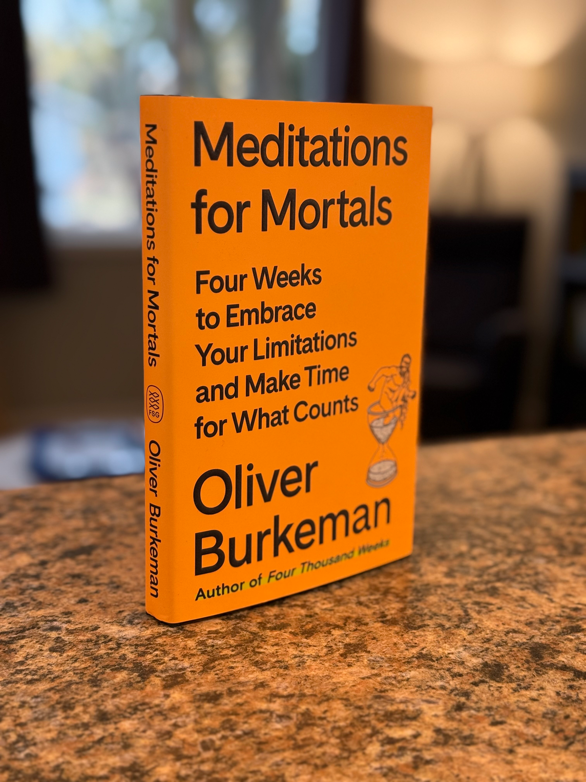 A hardcover copy of Meditations For Mortals by Oliver Burkeman stands on a granite countertop in a sunny room.