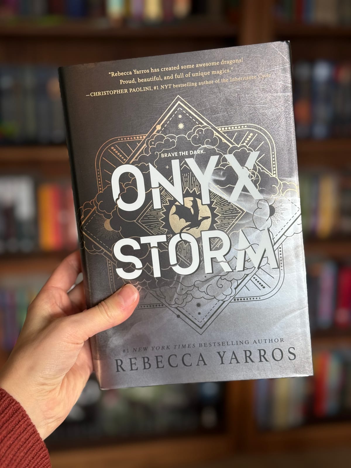 A hand holding up a hardcover copy of Onyx Storm by Rebecca Yarros in front of a stuffed bookcase.