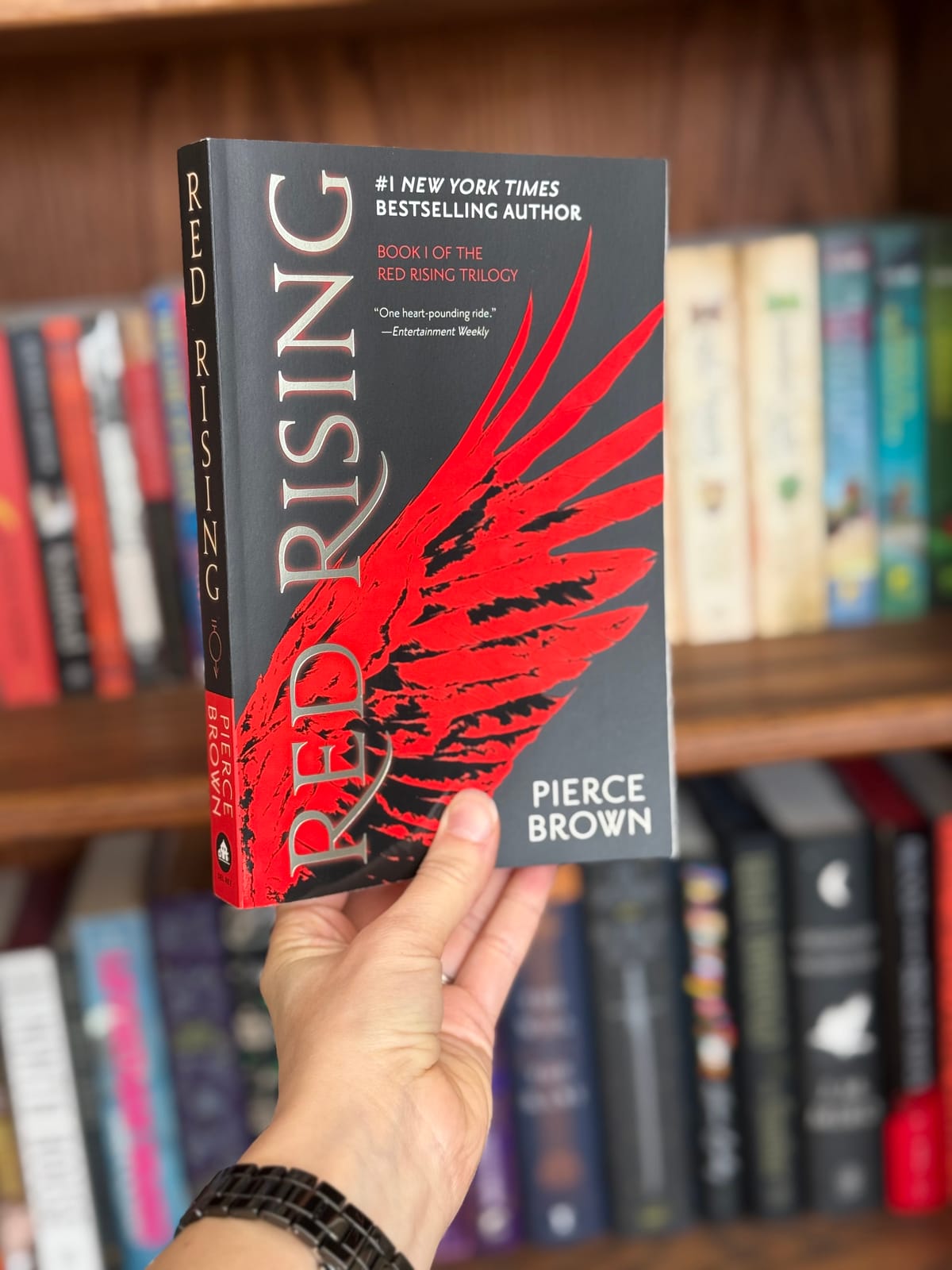 A woman holding a paperback copy of Red Rising by Pierce Brown in front of a stuffed bookcase.