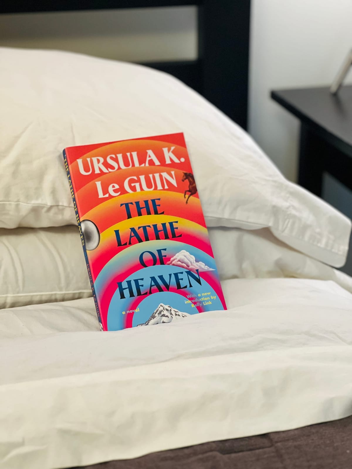 A paperback copy of The Lathe of Heaven by Ursula K. Le Guin is propped up against a white pillow on a neatly made bed.