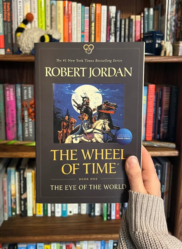 A woman's hand holding up a paperback copy of The Eye of The World by Robert Jordan in front of a stuffed bookcase.