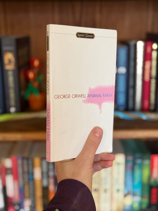 A hand holding a paperback copy of Animal Farm by George Orwell in front of a stuffed bookcase.