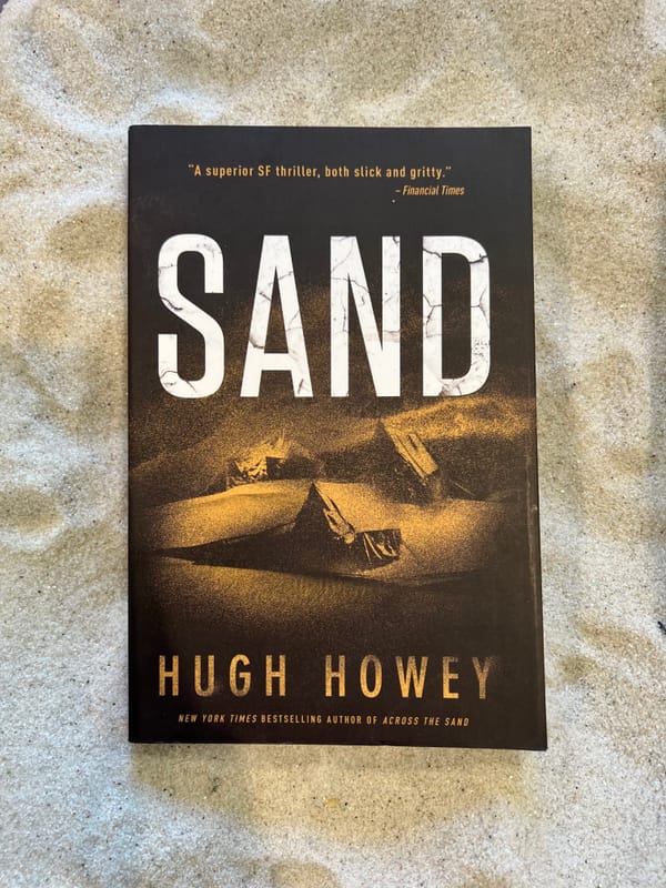 A paperback copy of Sand by Hugh Howey is lying in a bed of white sand.