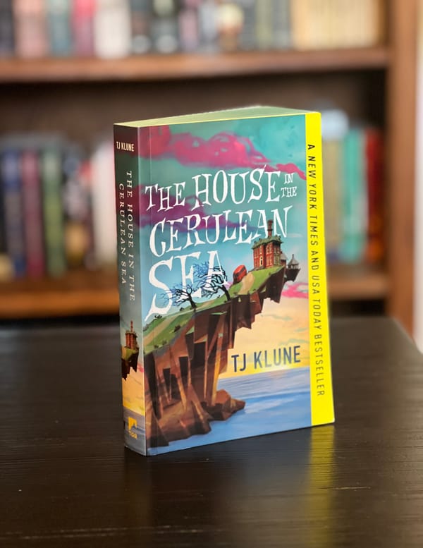 A paperback copy of The House In The Cerulean Sea by TJ Klune in front of a stuffed bookcase.