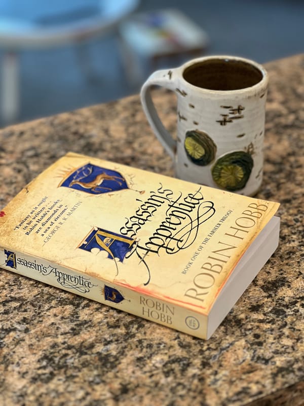 A paperback copy of Assassin's Apprentice by Robin Hobb lying on a granite countertop next to a cup of coffee.