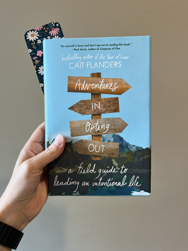 A hand holding up a hardcover copy of Adventures in Opting Out by Cait Flanders.