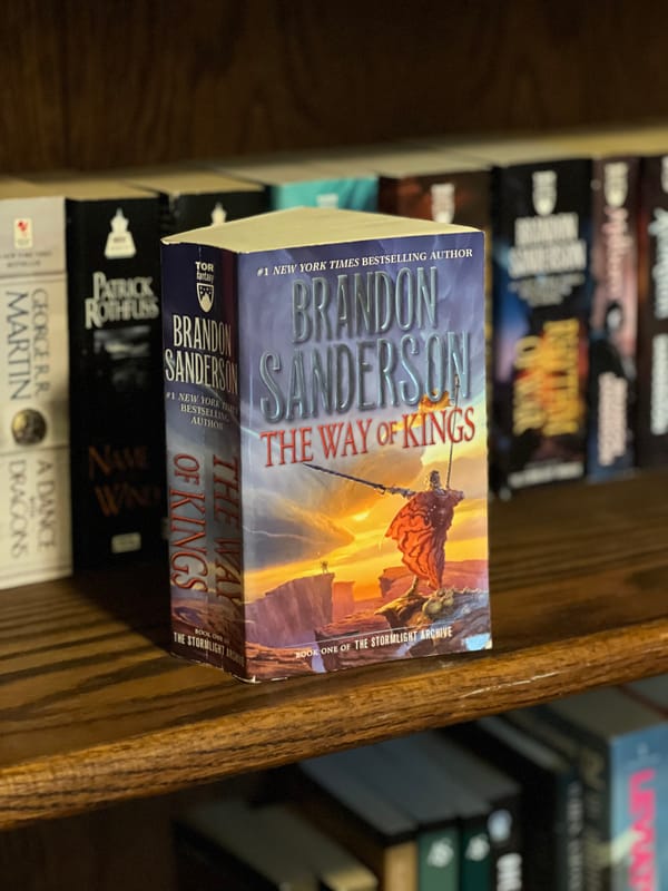 A paperback copy of The Way Of Kings by Brandon Sanderson stands on a bookshelf in front of a stuffed bookcase.