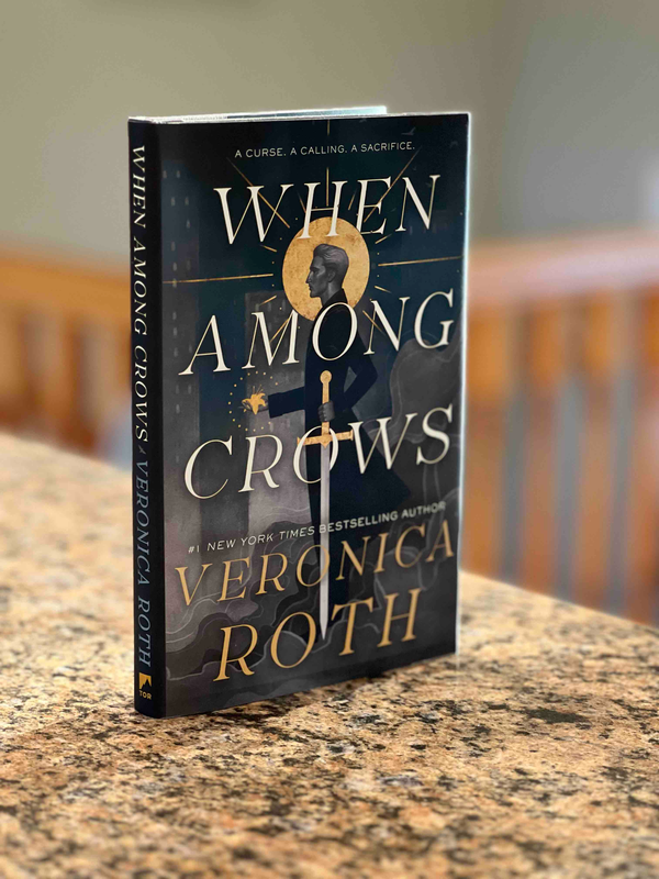 A hardcover copy of When Among Crows by Veronica Roth stands on a granite countertop in a sunny room.