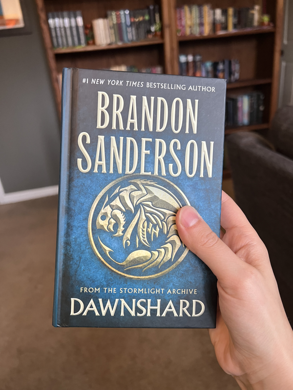 A hand holding a hardcover copy of Dawnshard by Brandon Sanderson in front of a stuffed bookcase.