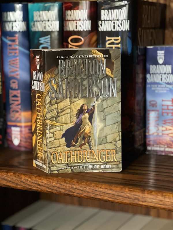 A paperback copy of Oathbringer by Brandon Sanderson stands on a shelf in front of a stuffed bookcase.